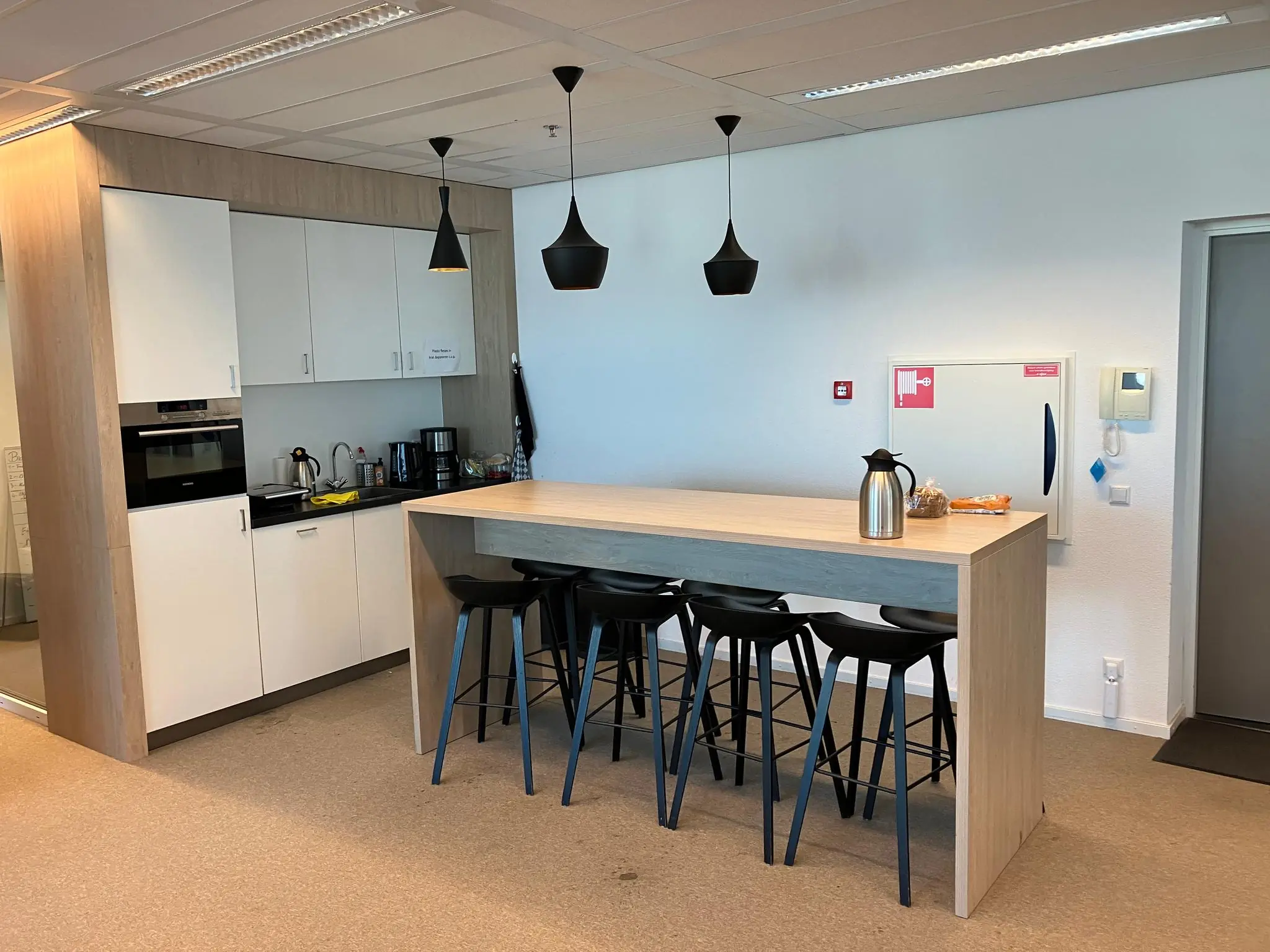 Moderne kantoorpantry met houten bar, zwarte barkrukken en koffiezetapparaten.